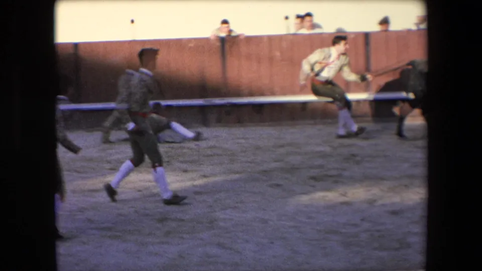 A dark bull moves across the Barcelona arena as a matador in a decorated jacket and several other performers react nearby in the sand ring.