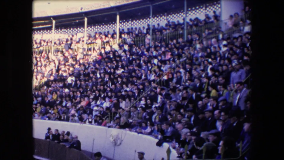 A crowded Barcelona stadium grandstand appears above the bullring wall, with many spectators seated and standing in bright sun and shadow.