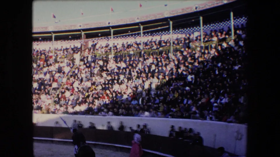 Wide 1971 Barcelona bullring view with sunlit upper seating, shadowed lower crowd areas, and a pink-costumed figure near the ring edge.