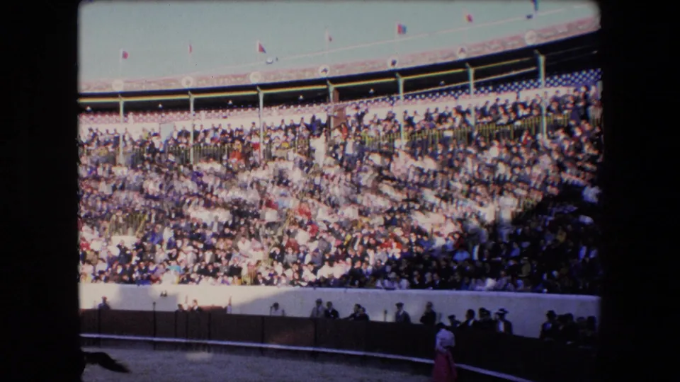 1971 Barcelona home-movie frame of a bullring audience, with small flags along the upper edge and people gathered near the lower arena barrier.