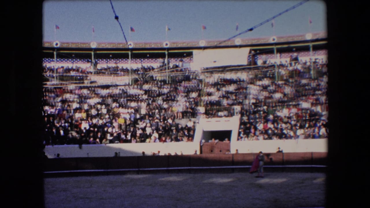 Wide 1971 Barcelona bullfighting arena view with tiered spectators, bright sun, flags above the stands, and a small matador holding a red cape near the sandy ring.