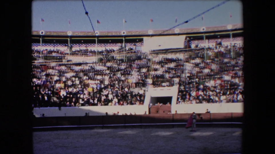 Wide 1971 Barcelona bullfighting arena view with tiered spectators, bright sun, flags above the stands, and a small matador holding a red cape near the sandy ring.