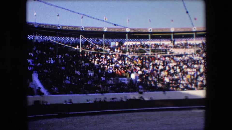 Visitor-shot 8mm frame from a 1971 Barcelona bullfighting arena showing crowded stands, shaded seating, overhead lines, and the sandy ring foreground.