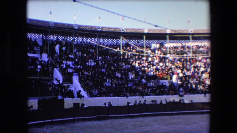 Wide 1971 Barcelona bullring frame with spectators packed into tiered seating, a stairway at left, flags overhead, and the sandy arena edge below.