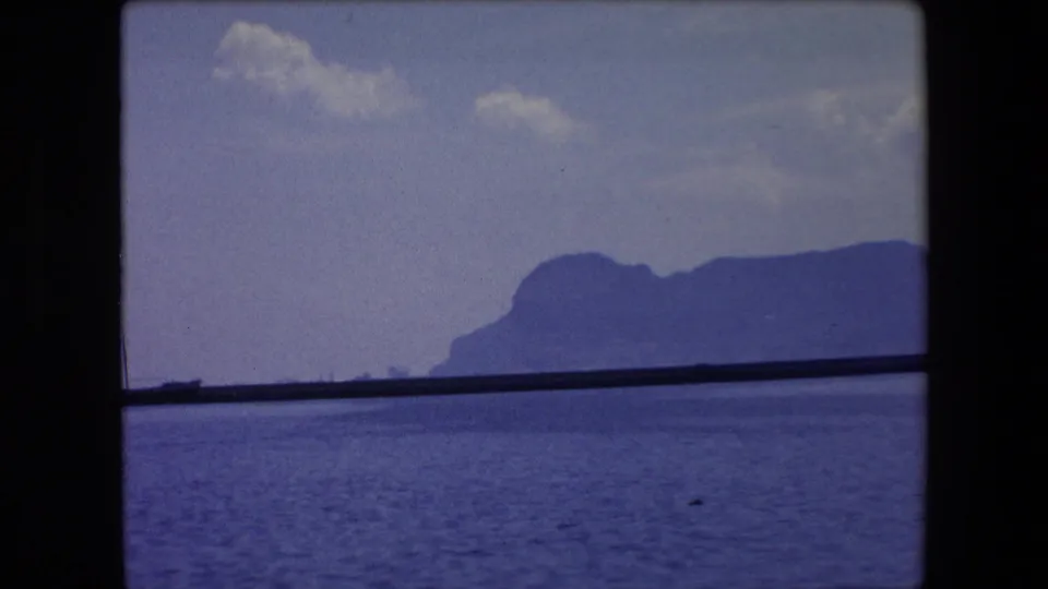 The clip cuts to a coastal waterside view in the supplied Barcelona travel footage, with calm water, a dark shoreline, small boats, and a distant mountain silhouette.