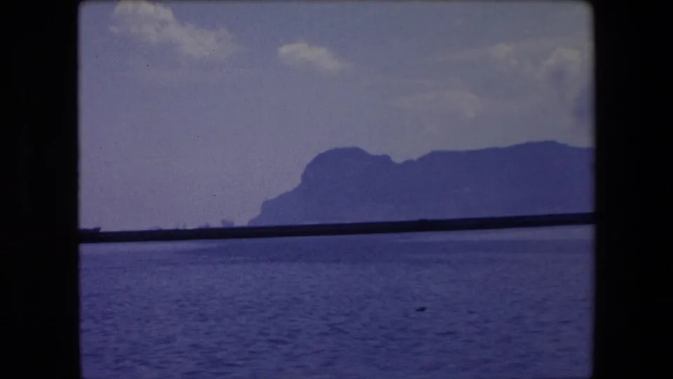 A second coastal view from the 1971 Barcelona home movie shows blue-toned water, a distant mountain ridge, faint boats, and pale cloud cover.