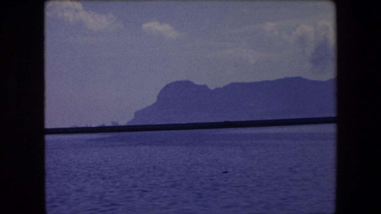 1971 Barcelona-area home-movie frame showing calm sea, a distant mountainous coastline, and a dark horizontal window or rail crossing the view.