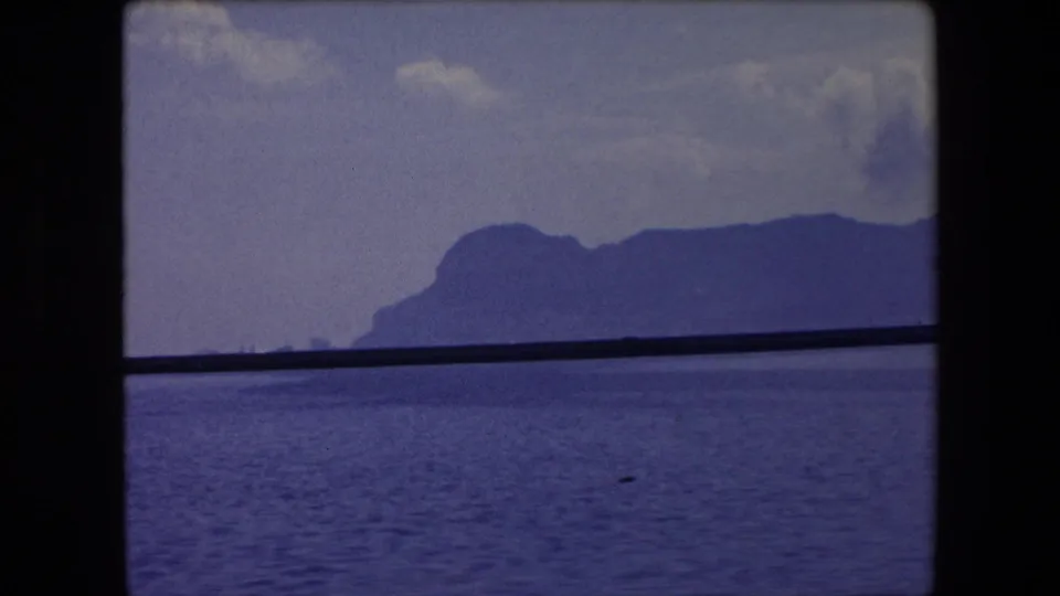 1971 Barcelona-area home-movie frame showing calm sea, a distant mountainous coastline, and a dark horizontal window or rail crossing the view.
