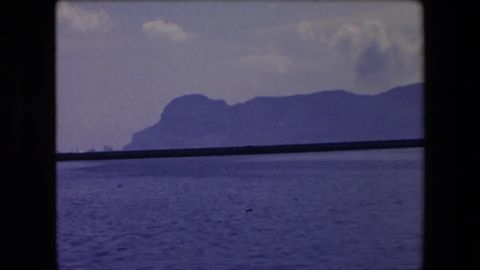 1971 Barcelona-area seascape with open water in the foreground, a dark mid-frame rail or window bar, and hazy mountains along the horizon.
