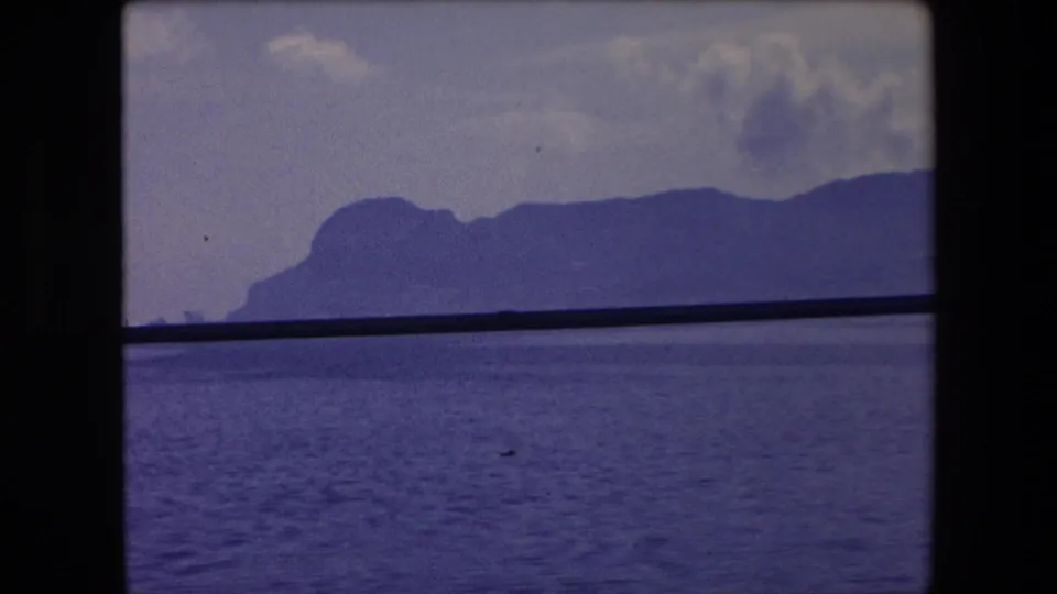 8mm home-movie frame from the 1971 Barcelona clip showing rippled sea, distant coastal mountains, and a dark rail or window edge across the middle ground.