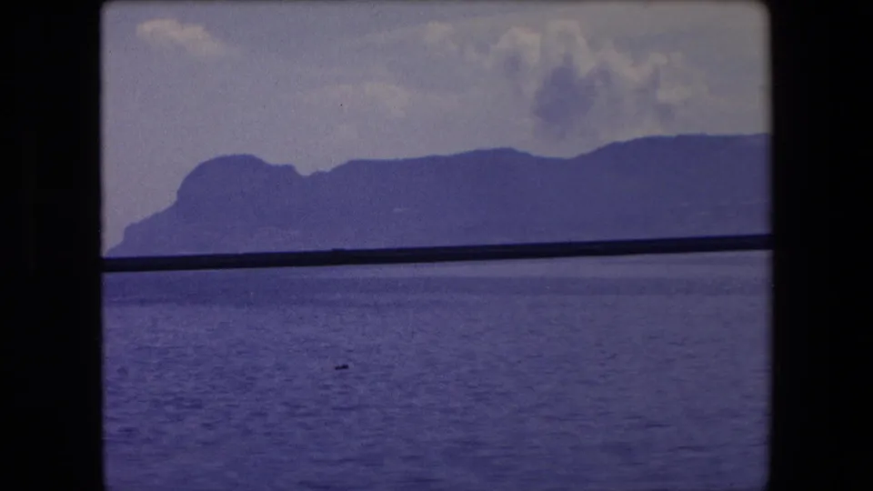 Final frame of the 1971 Barcelona-area seascape, with calm water, hazy coastal hills, and the dark boat-window or rail element still crossing the view.
