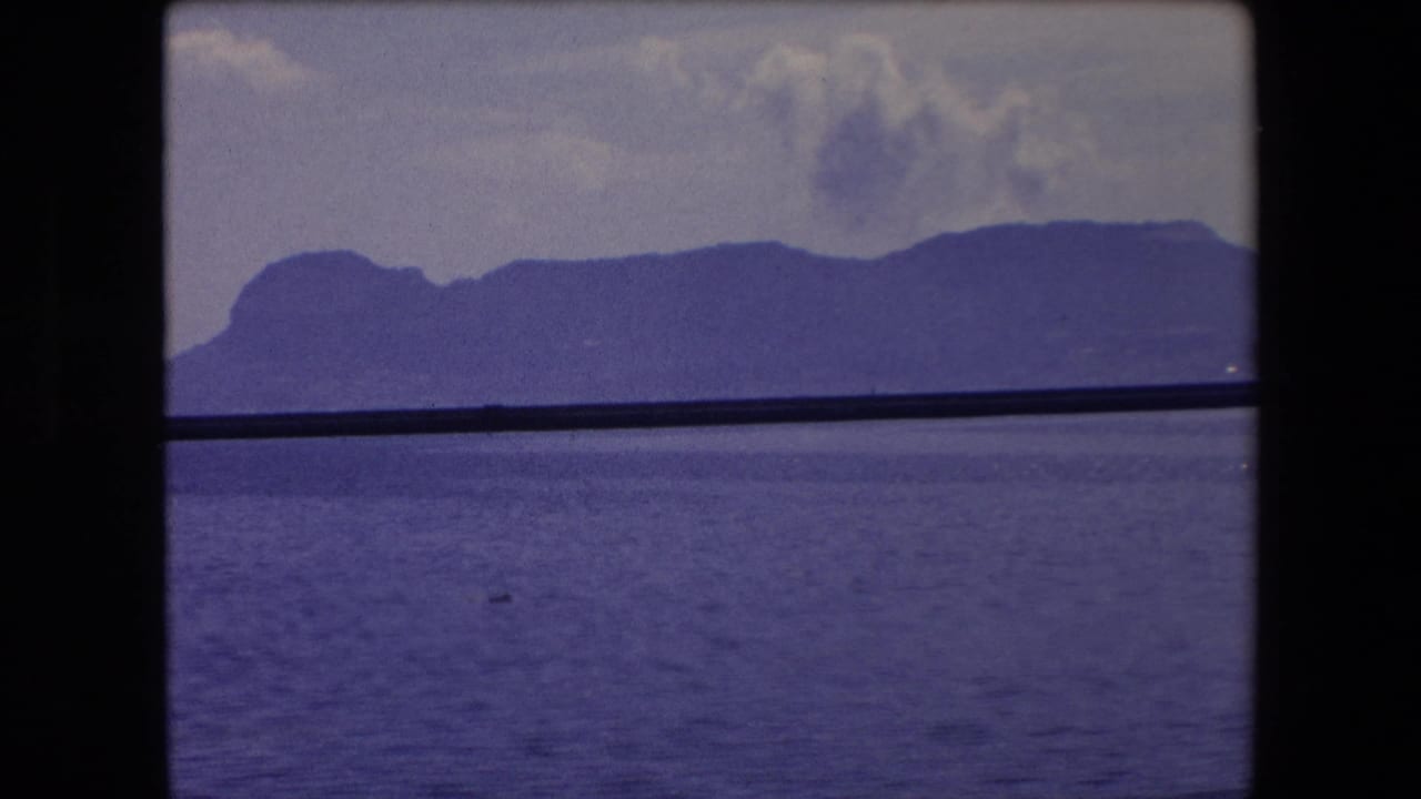 1971 Barcelona-area 8mm frame showing a distant mountain ridge over blue water, with a dark vertical divider and horizontal bar crossing the view.