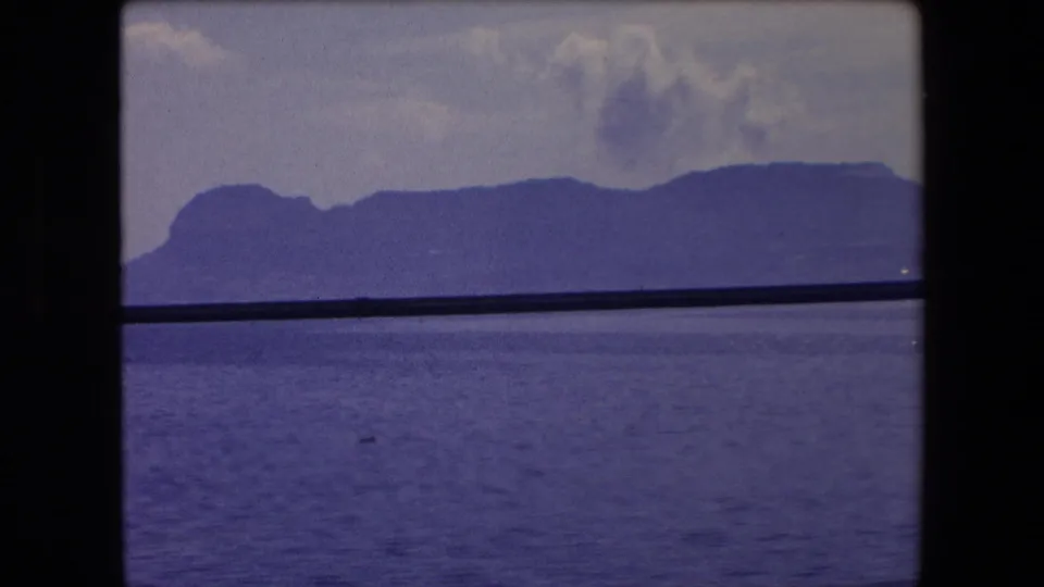 1971 Barcelona-area 8mm frame showing a distant mountain ridge over blue water, with a dark vertical divider and horizontal bar crossing the view.
