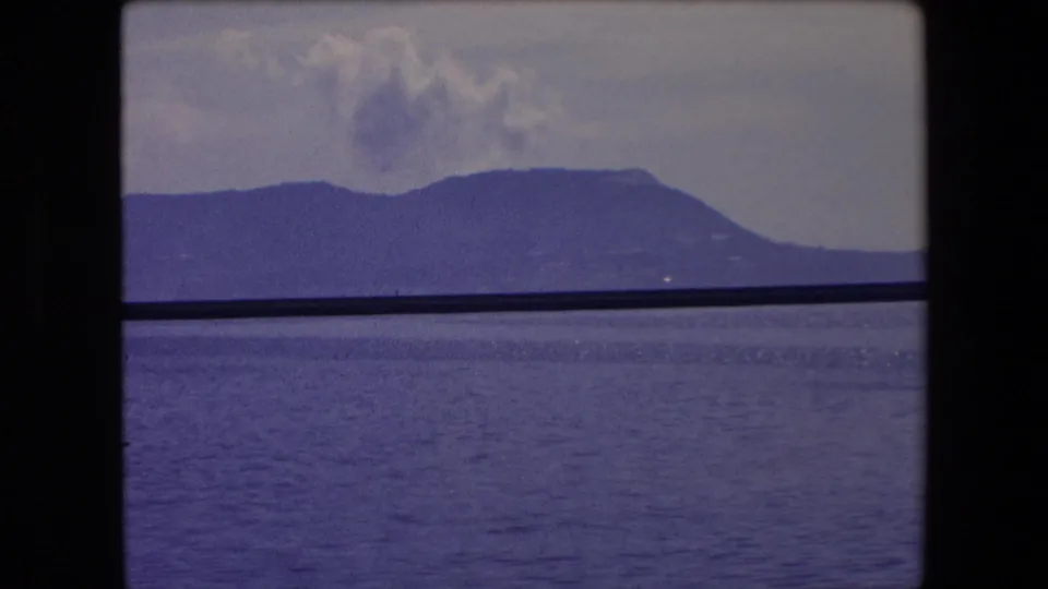 1971 Barcelona-area 8mm frame showing water in the foreground, a distant mountain slope, and soft cloud forms, with dark frame bars at the edges.
