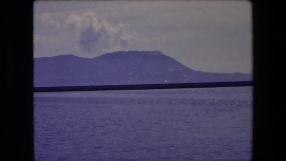 1971 Barcelona-area 8mm frame of blue water, a dark mountain ridge, and smoke-like clouds above, partially obscured by a horizontal bar.