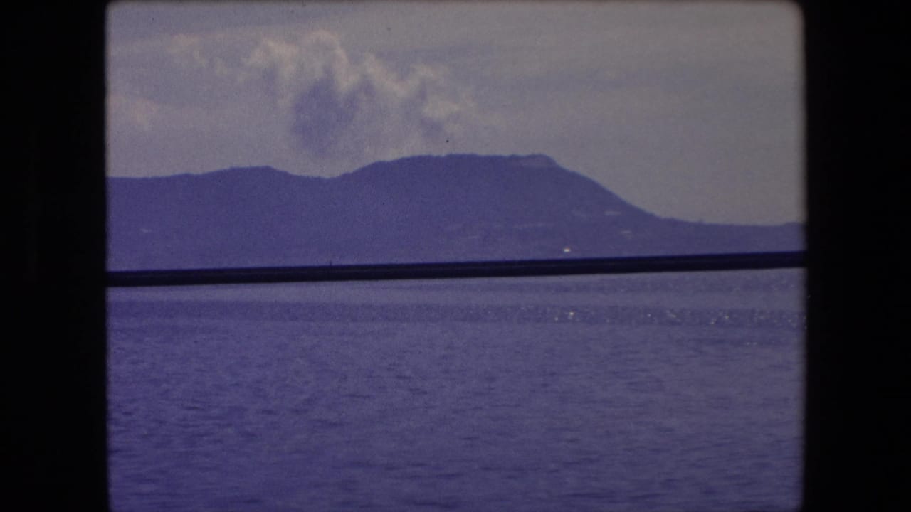 1971 Barcelona-area coastal view with blue water, a distant mountain ridge, pale clouds, and a dark horizontal edge crossing the frame.