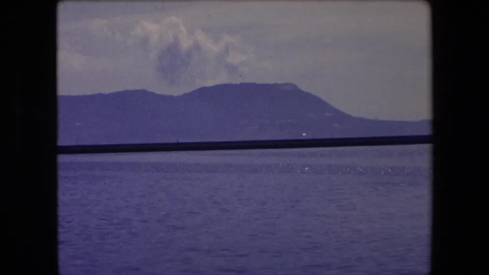 1971 Barcelona-area coastal view with blue water, a distant mountain ridge, pale clouds, and a dark horizontal edge crossing the frame.