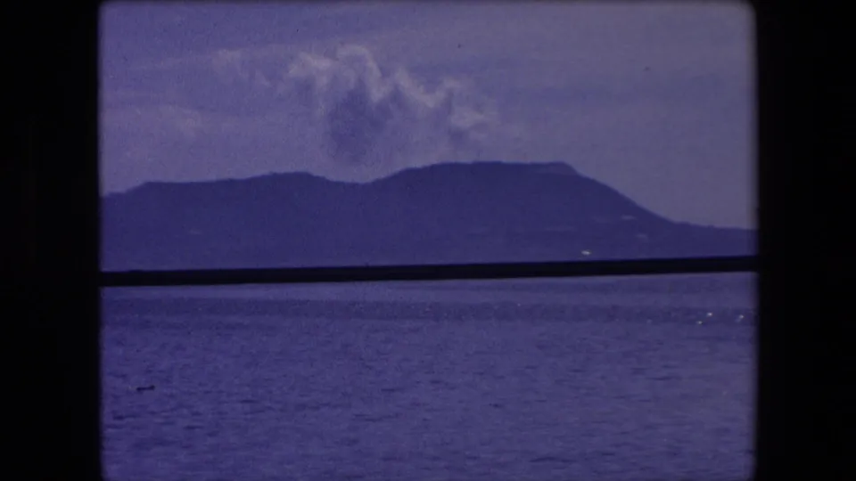 A blue-toned 8mm frame shows distant Spanish coastal mountains across the water, with low clouds and a dark railing or window edge bisecting the view.