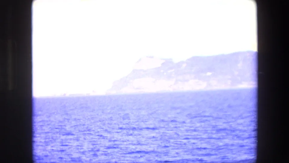 Overexposed 8mm travel frame showing bright sea in the foreground and a pale coastal mountain mass partly washed out by strong light.