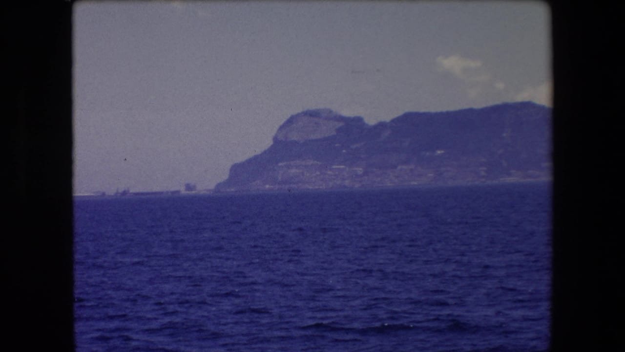 Blue Mediterranean water fills the lower frame while a distant rocky shoreline near Barcelona appears through haze beneath a pale sky.