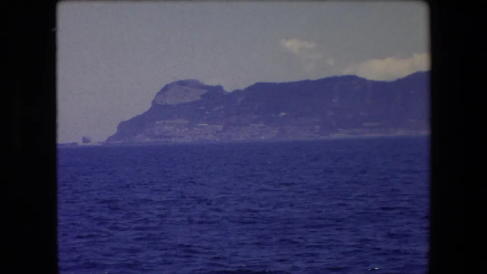 A steady 1971 travel shot frames blue sea below a dark, distant coastal ridge with pale clouds overhead near Barcelona, Spain.