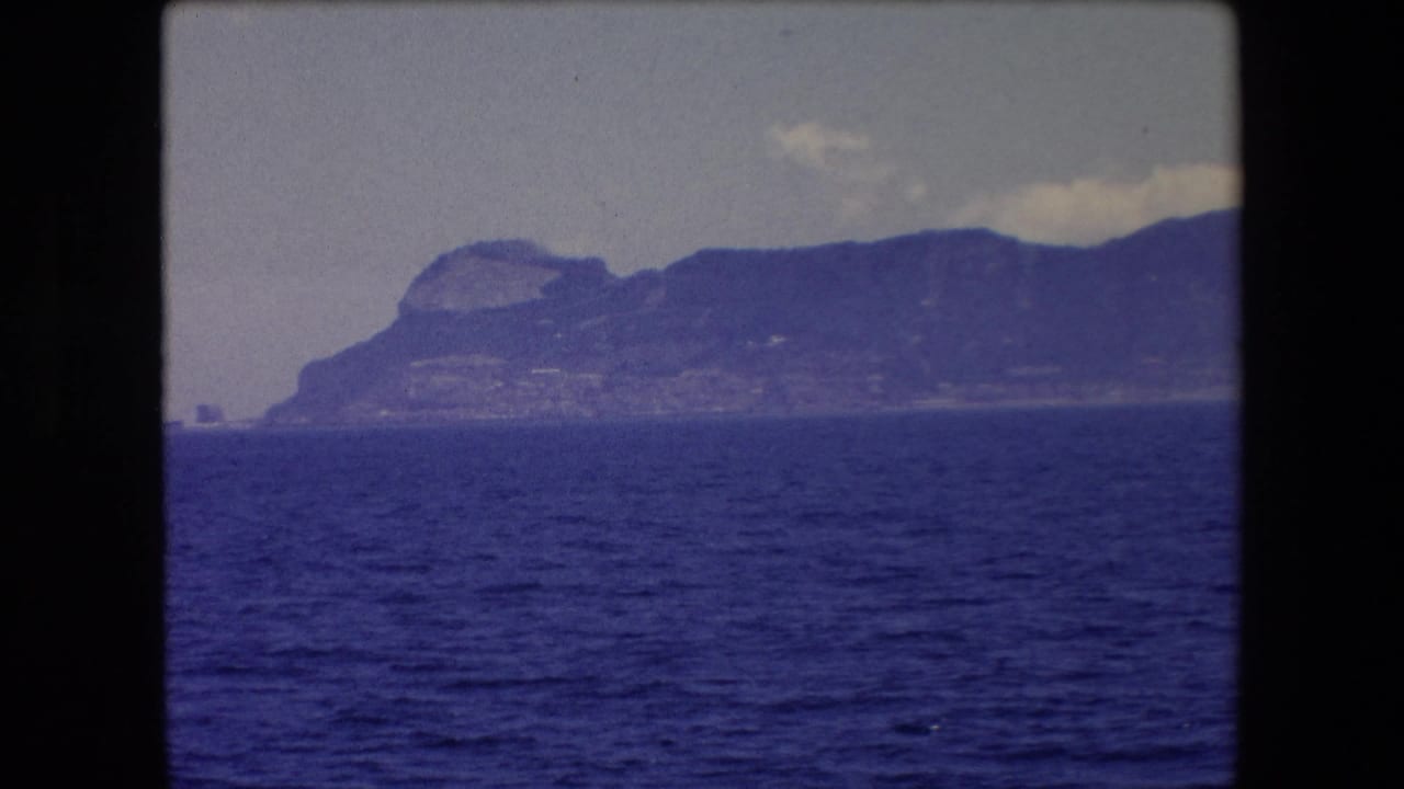 1971 8mm travel footage from Barcelona, Spain, showing a hazy mountainous coastline viewed across deep blue water beneath a pale sky.