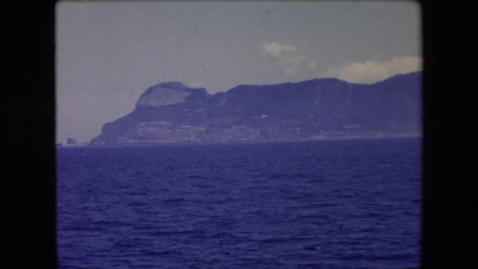 1971 8mm travel footage from Barcelona, Spain, showing a hazy mountainous coastline viewed across deep blue water beneath a pale sky.
