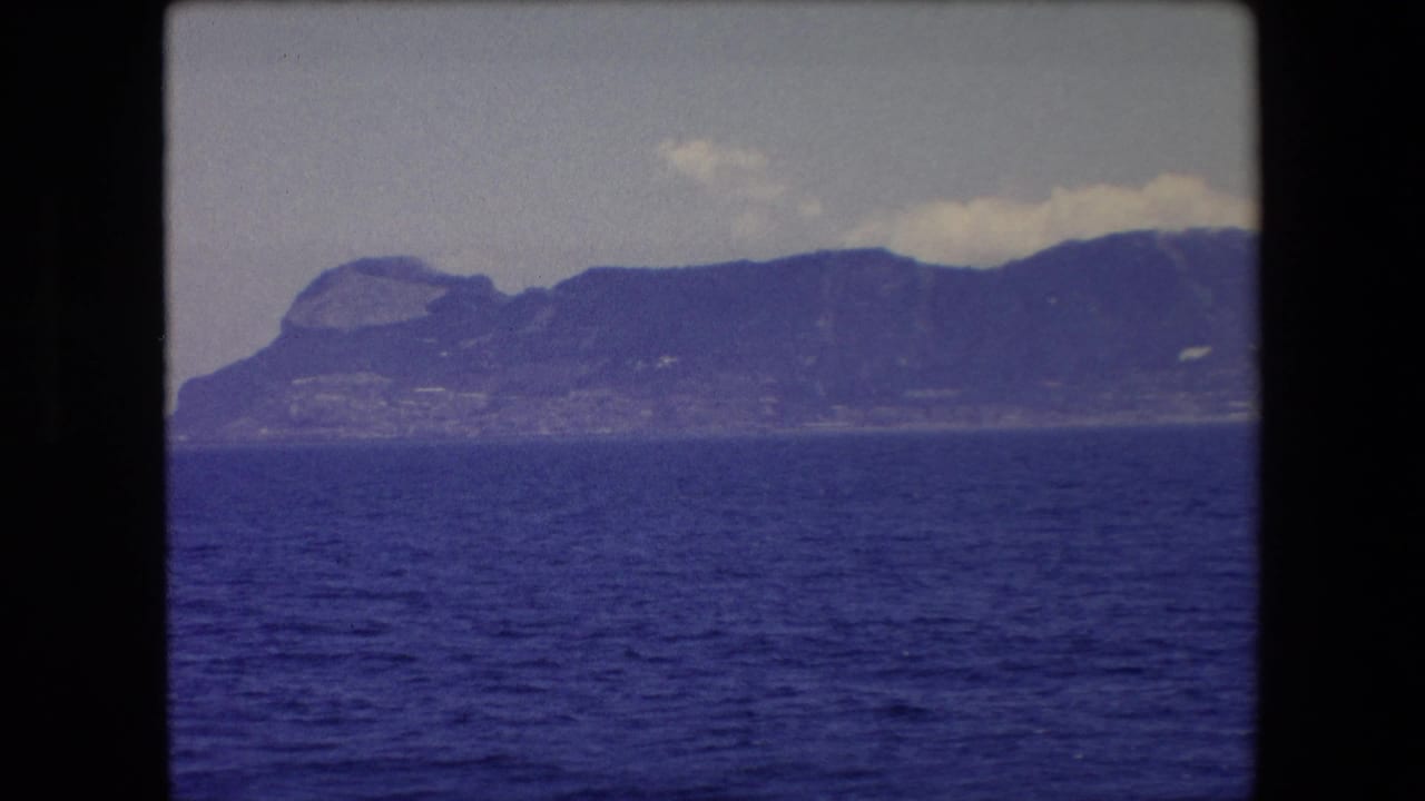 1971 8mm Barcelona-area seascape with rippling blue water in the foreground and a rugged mountainous shoreline in haze.