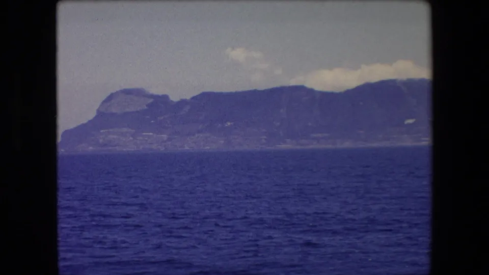 1971 8mm Barcelona-area seascape with rippling blue water in the foreground and a rugged mountainous shoreline in haze.