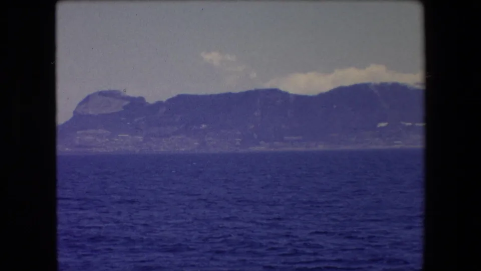 Soft 8mm frame from 1971 Barcelona travel footage showing blue sea, a hazy distant shoreline, and a dark mountain ridge under thin clouds.