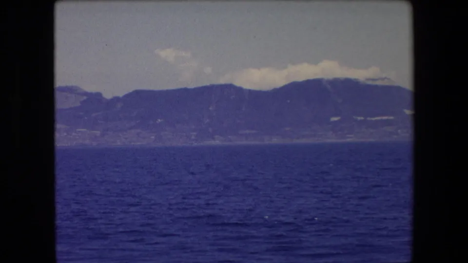 1971 8mm seascape near Barcelona, Spain, with open blue water, a distant mountainous coastline, and pale clouds along the horizon.