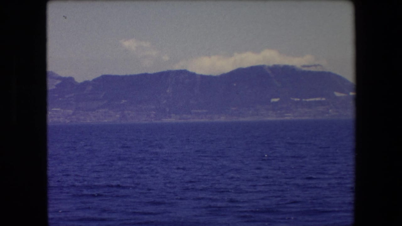 1971 Barcelona-area home-movie frame showing a distant mountainous shoreline across deep blue water under a pale cloudy sky.