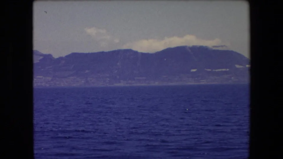 1971 Barcelona-area home-movie frame showing a distant mountainous shoreline across deep blue water under a pale cloudy sky.