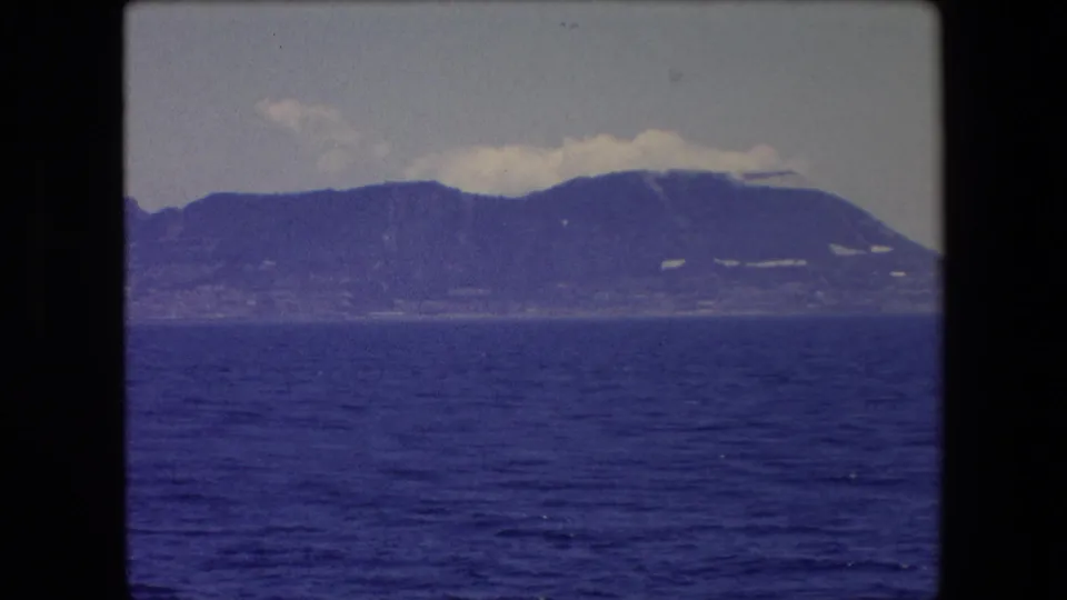 1971 Spain travel-film view of a rugged coastal landmass seen from across the water, with saturated blue sea and soft distant detail.