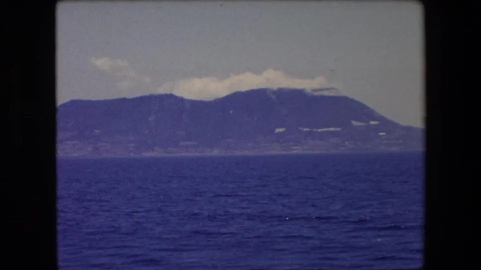 1971 Barcelona coastal home-movie frame with open water in the foreground and a hazy mountain ridge along the distant shore.