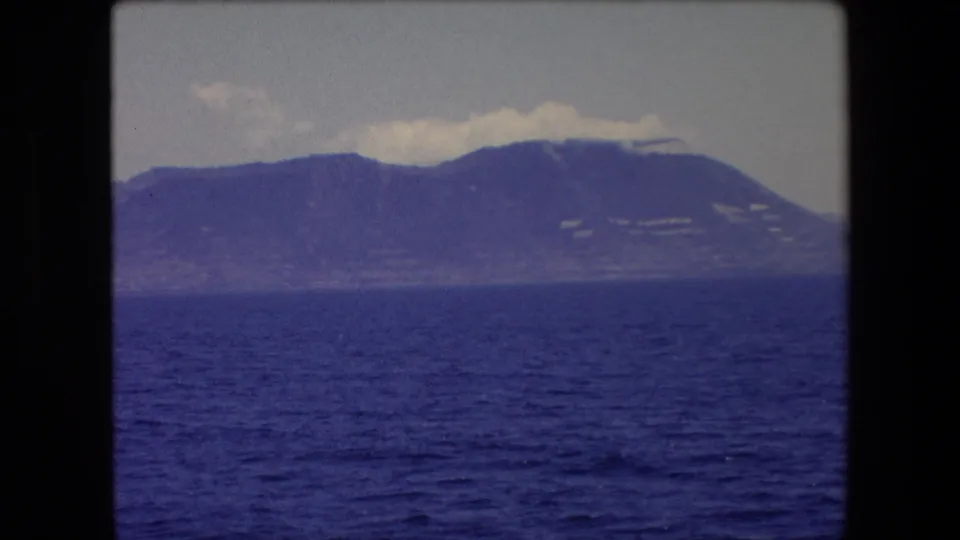 1971 Spain 8mm frame showing a blue sea foreground and distant coastal mountains, with slight haze and visible amateur-film texture.