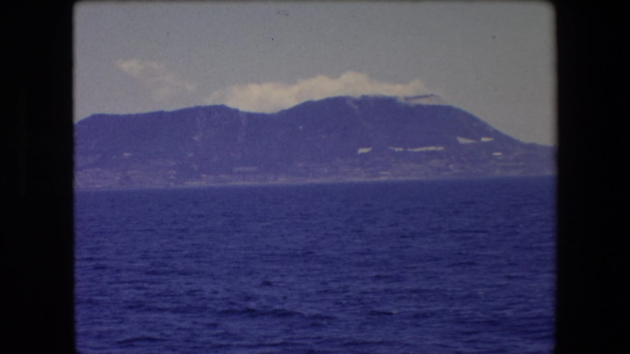 1971 Barcelona home-movie frame showing dark blue sea in the foreground with a hazy distant mountain ridge and low clouds over the peaks.