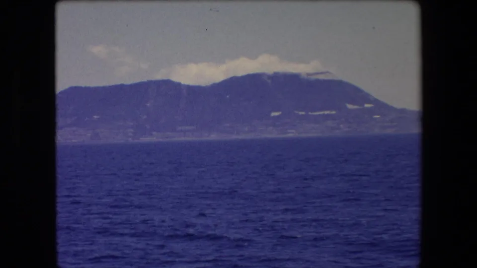 1971 Barcelona home-movie frame showing dark blue sea in the foreground with a hazy distant mountain ridge and low clouds over the peaks.