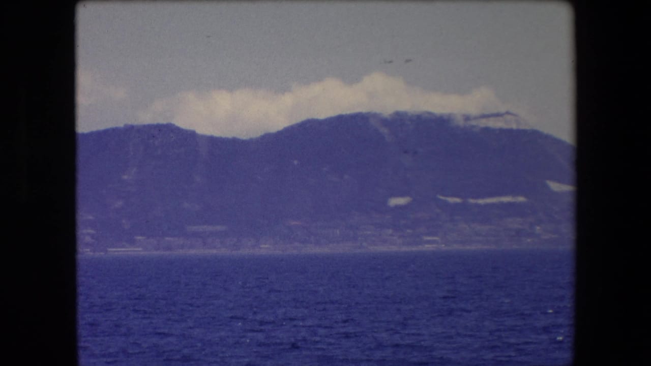 Mountain ridge and shoreline seen across blue water with clouds resting along the peaks.