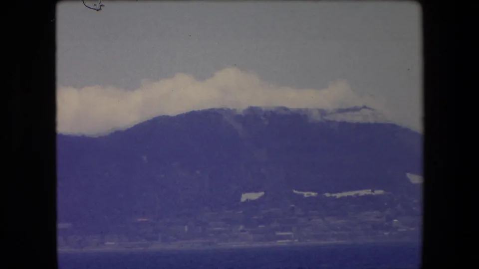 1971 Barcelona 8mm frame with the distant mountain range filling more of the view, clouds along the crest, and a narrow coastal strip near the waterline.