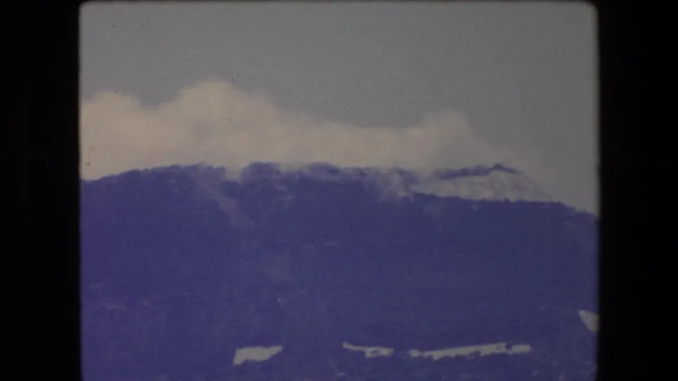 1971 Barcelona landscape frame showing a soft, purple-blue mountain ridge under low clouds, with only a narrow band of sea visible at the bottom.
