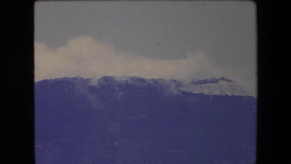 1971 8mm travel footage from the Barcelona area showing a distant mountain ridge under a cloudy sky, with pale snow or exposed rock near the upper slope.