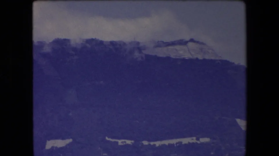 1971 home-movie frame showing a shadowed mountain ridge under clouds, with indistinct pale foreground shapes along the bottom of the frame.