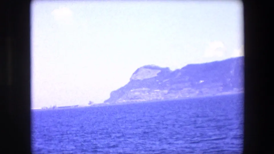 1971 8mm travel view from the Barcelona area looking across blue sea toward a rocky mountainous coastline under a bright, overexposed sky.