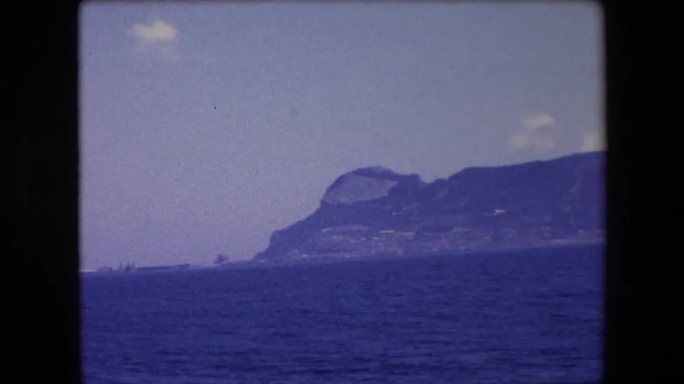 1971 8mm home-movie frame from Barcelona, Spain, showing a distant rugged cliff formation viewed across the Mediterranean from a vessel.