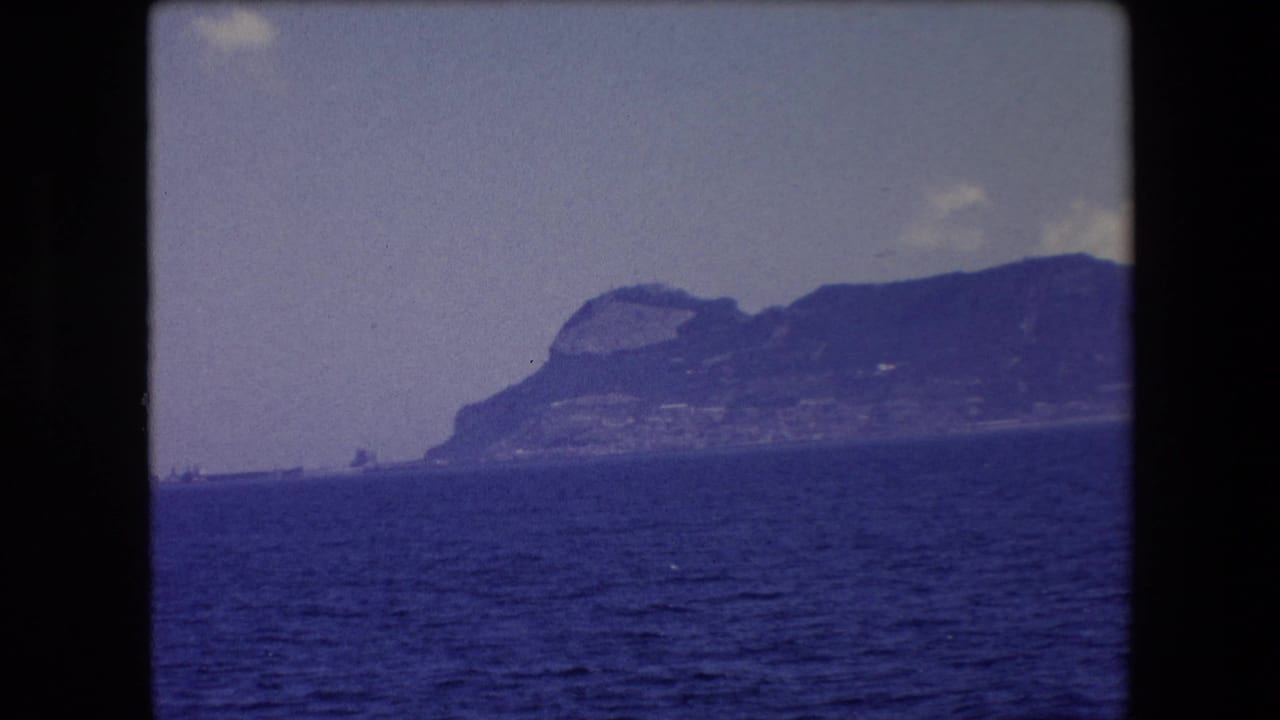 A soft 8mm frame shows the Barcelona-area coastline in 1971, with a rocky headland rising behind the sea and small cloud forms above it.