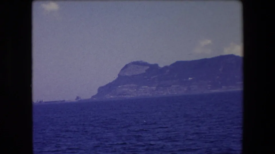 A soft 8mm frame shows the Barcelona-area coastline in 1971, with a rocky headland rising behind the sea and small cloud forms above it.