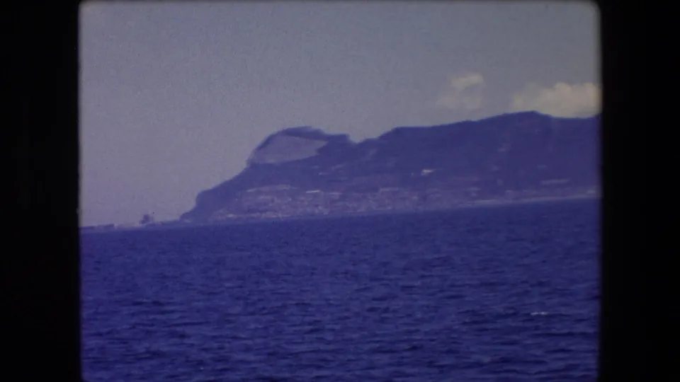 1971 8mm travel footage frame from Barcelona, Spain, showing open Mediterranean water in the foreground and a distant cliff-backed coastline under a hazy sky.