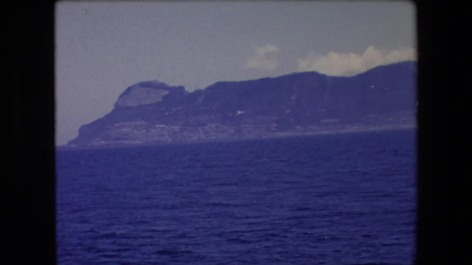 The final 1971 8mm frame holds on the Barcelona-area coastal ridge, with textured blue water, a pale sky, and scattered clouds above the distant rock formation.