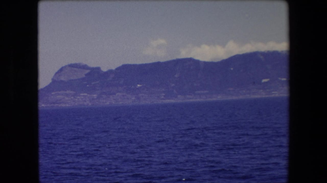 1971 Barcelona-area 8mm travel frame showing a distant mountainous coast or island beyond dark blue water, with pale clouds above and dark film borders around the frame.